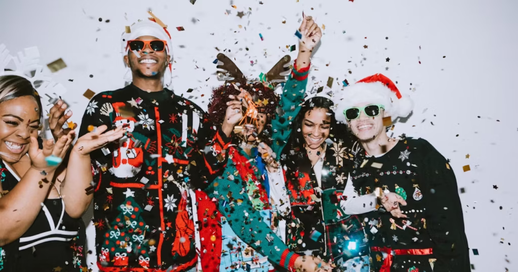 A group of friends wearing Christmas sweaters and festive accessories celebrate together, smiling and laughing as colorful confetti falls around them at a holiday party.