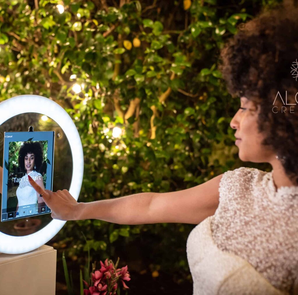 Woman interacting with a touchscreen mirror photo booth in a lush outdoor setting, illuminated by soft lights, showcasing modern wedding entertainment in Charlotte.