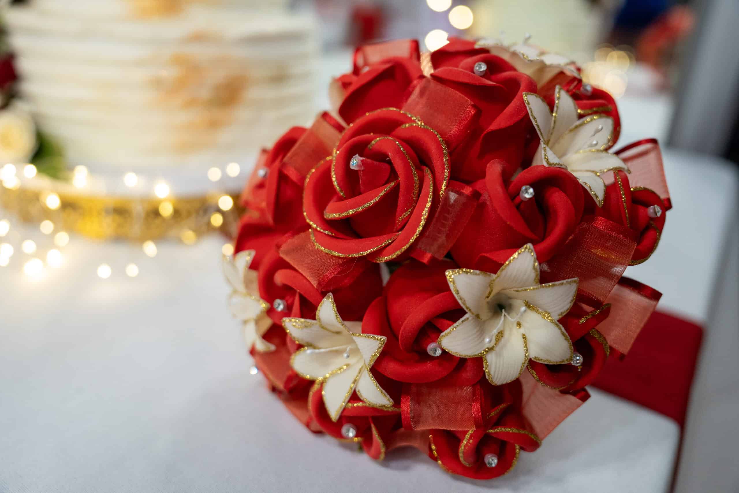 Bouquet of red fabric roses and white flowers with gold accents, set against a blurred background of a wedding cake and twinkling lights, symbolizing event elegance and celebration.
