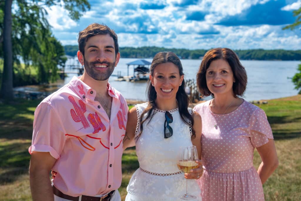 Group of three smiling individuals at a lakeside event, with the woman in a white dress holding a glass of wine, surrounded by lush greenery and a scenic water view, capturing a joyful moment that embodies the essence of celebrations and gatherings.