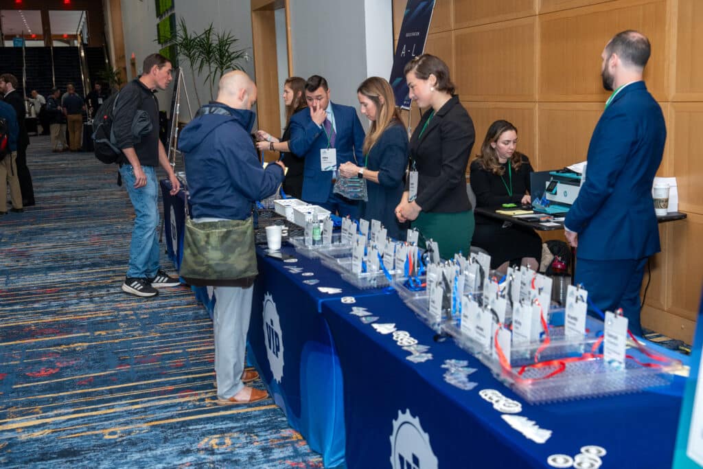 Event attendees engaging at a registration table, featuring a blue tablecloth displaying event materials and promotional items, highlighting corporate networking and professional interactions at a conference.