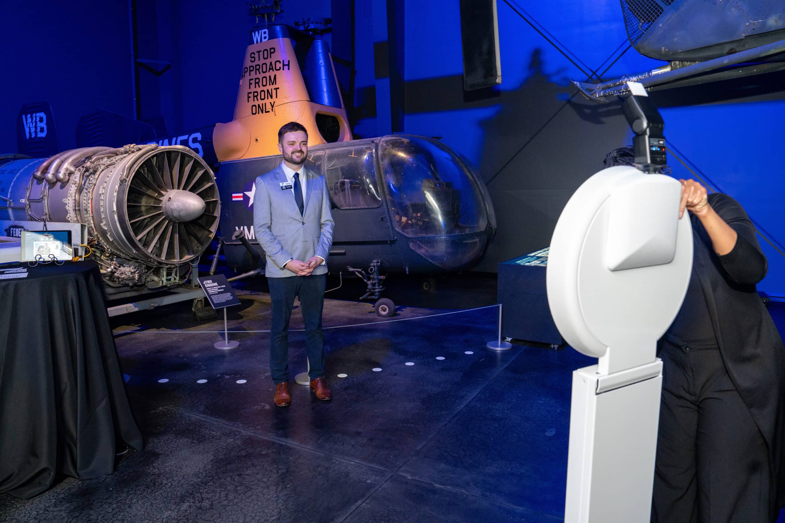 Event staff member engaging with guests at an aviation-themed event, featuring a helicopter and jet engine display in a blue-lit venue.