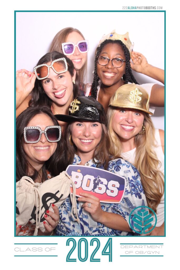 Group of six women posing in a photo booth, wearing fun accessories like oversized sunglasses and hats, holding props including a sign that says "BOSS" and a money bag, celebrating the Class of 2024 for the Department of OB/GYN.