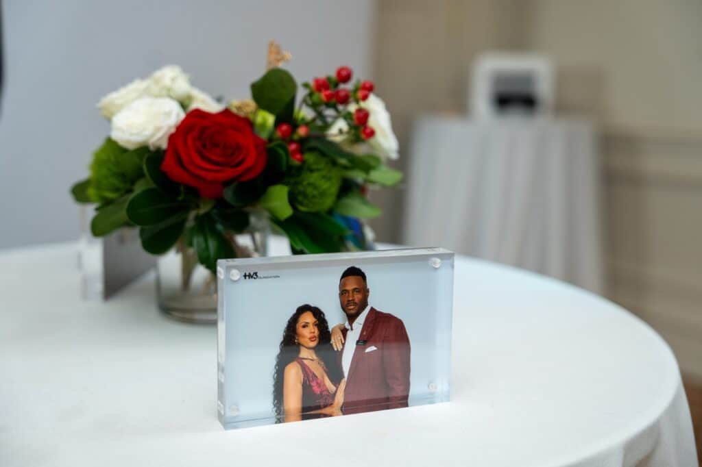 A glam booth portrait of a couple displayed in a clear acrylic frame on a white table beside red and white flowers
