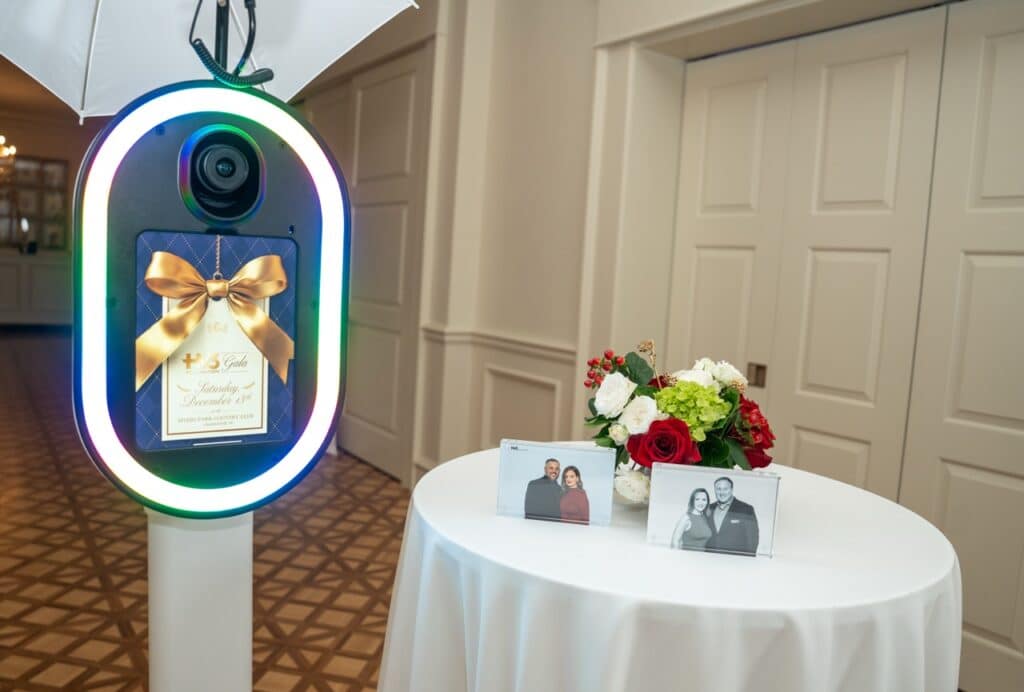 A glam photo booth displaying a custom gold-bow gala start screen, beside a table holding two black-and-white framed prints and red and white flowers